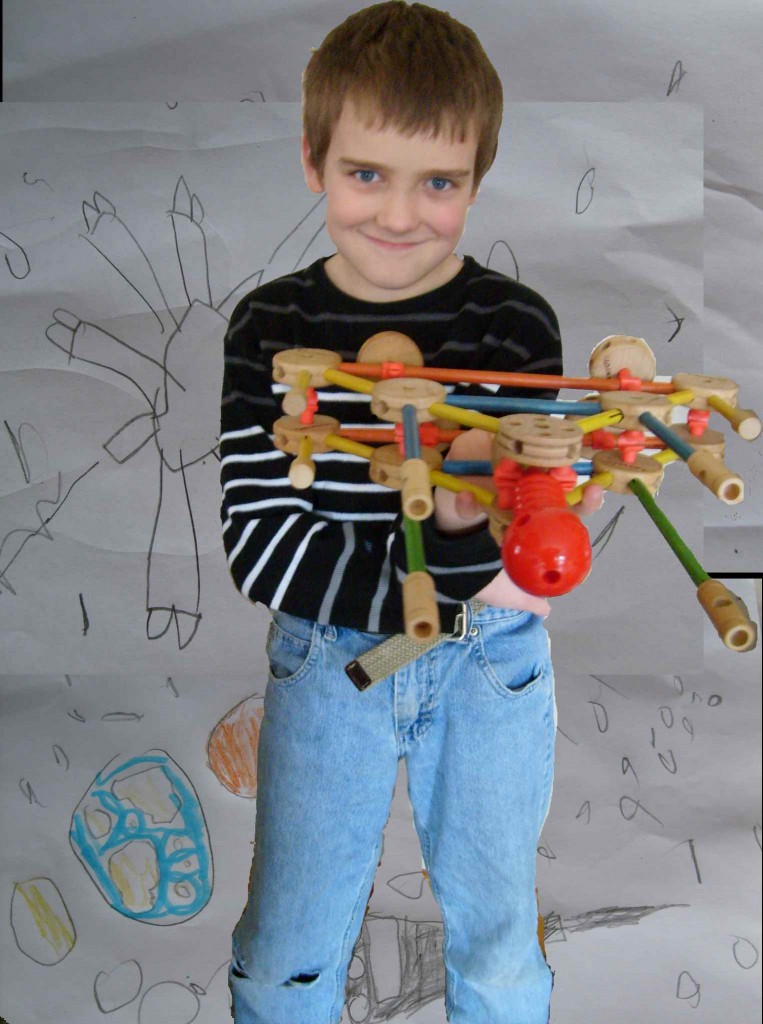 M holding a tinkertoy rocket ship with a background of his space drawings